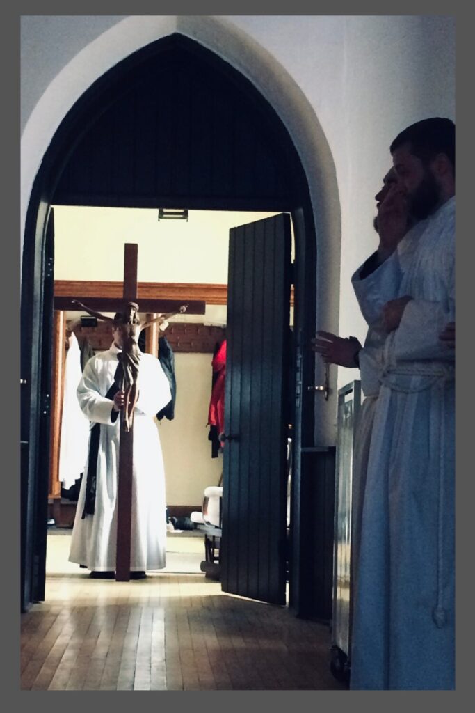 A man in a white chasable hods a large crucifix in front of his face as he enters a chapel, while other white-clad men stand waiting along the wall.