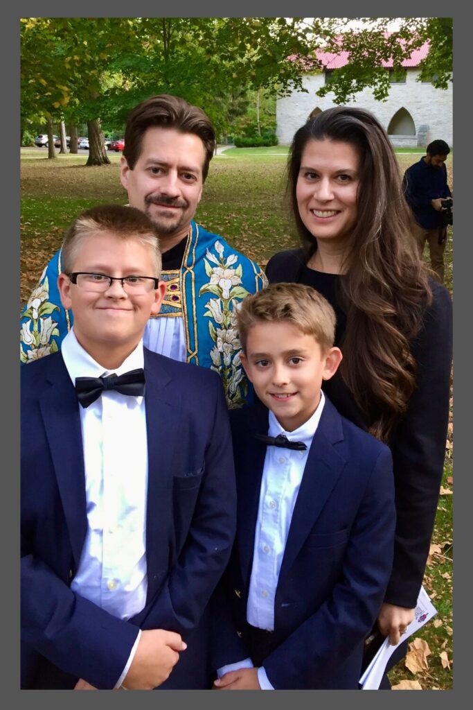 A priest in blue flowered vestments stand with his wife (the author of the article), older son with glasses, and younger son, both in bow ties, white shirts, and suit coats.