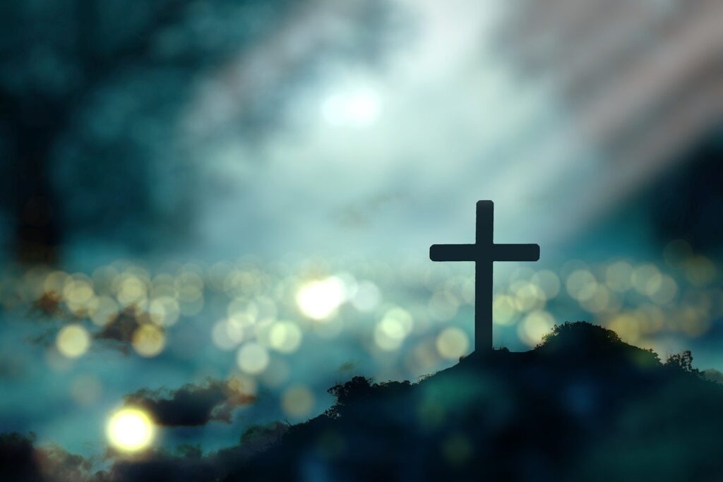 A small, thin wooden cross sits on the ground with sunlight in the background.