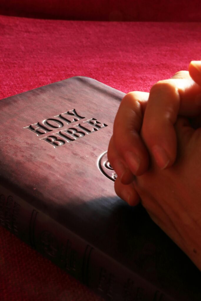 Hands are clasped together on a Christian Bible against a red background.
