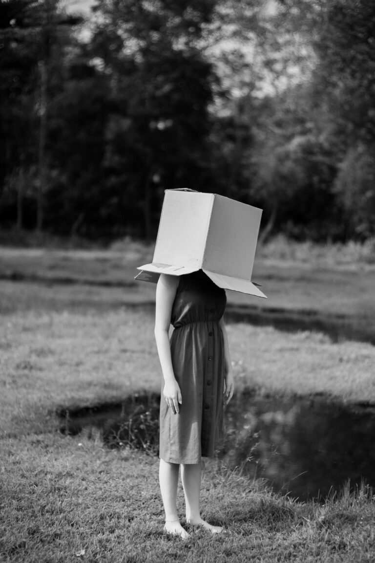 A black and white photo of a female standing with a box over her head.