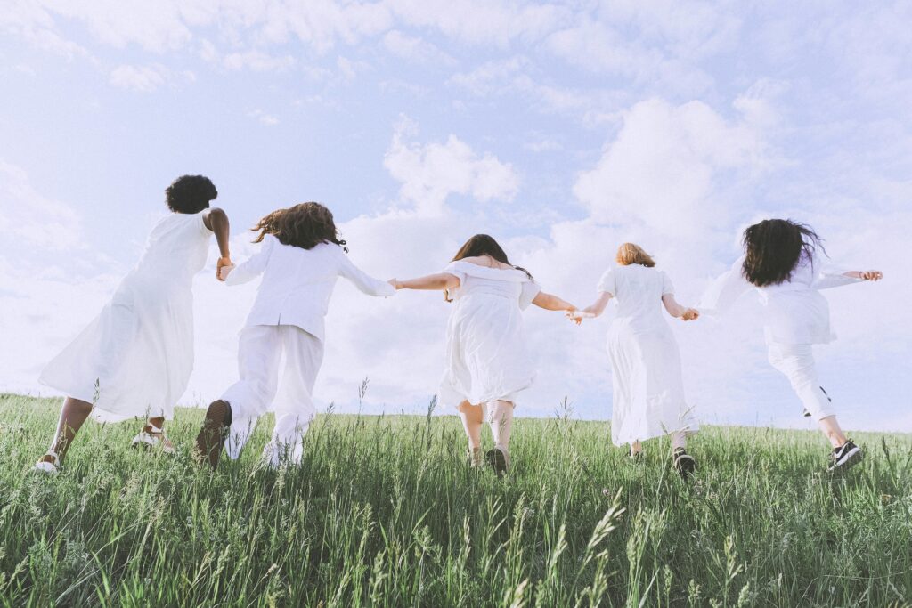 A line of five kids in white join hands and run with their backs to the camera on green grass against a blue sky.