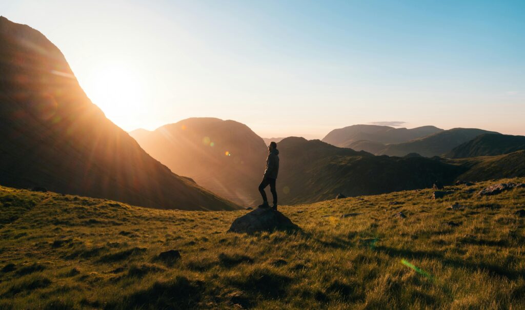 The silhouette of a person is almost illuminated by a sunrise peeking out from behind tall hills. 
